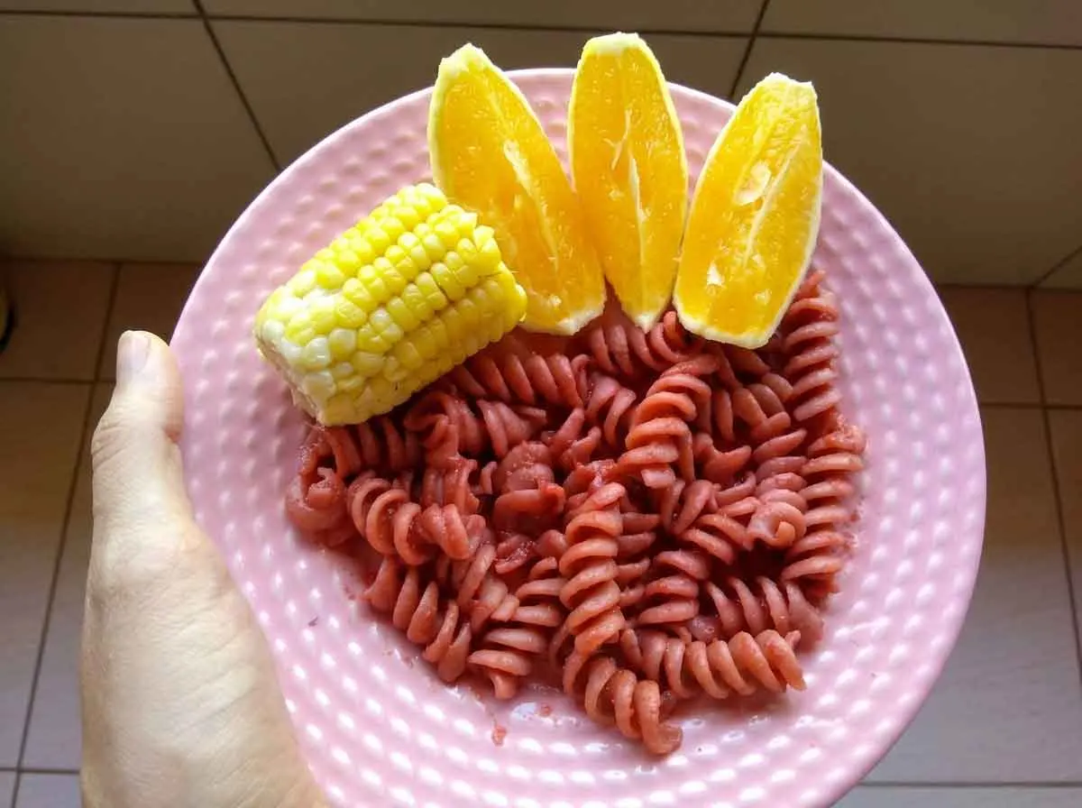 Prato colorido para criança com macarrão parafuso, milho na espiga e gomos de laranja